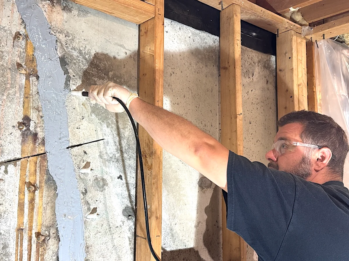 Technician performing polyurethane crack injection on a poured concrete foundation wall using injection packers and electric pumps.